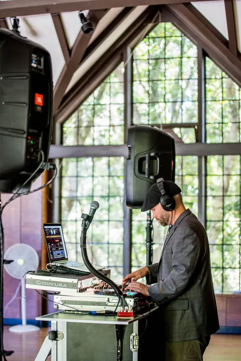 DJ Wayne Noel in headphones and a flat cap mixing in front of tall arched windows at the Brazilian Room in Tilden Park, Berkeley