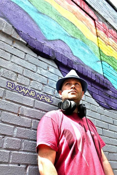 DJ Wayne Noel in a fedora and headphones against a painted brick wall in Brooklyn, New York