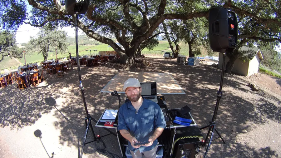 DJ Wayne Noel looking up from his DJ booth under a large oak tree at Kunde Family Winery in Kenwood, with speakers on stands and vineyard hills behind