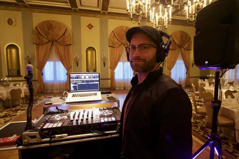 DJ Wayne Noel in headphones and a flat cap beside his DJ setup in the chandeliered ballroom at the Marines' Memorial Club in San Francisco