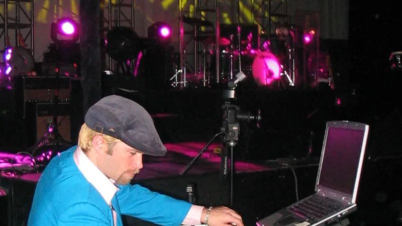 DJ Wayne Noel in a blue jacket and flat cap at his mixer and laptop in front of a concert stage, with a drum kit and magenta stage lights behind him, the night he opened for the B-52's