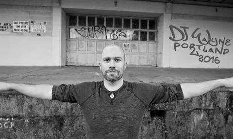Black and white portrait of DJ Wayne Noel, arms spread, in front of a graffiti wall that reads DJ Wayne Portland 2016
