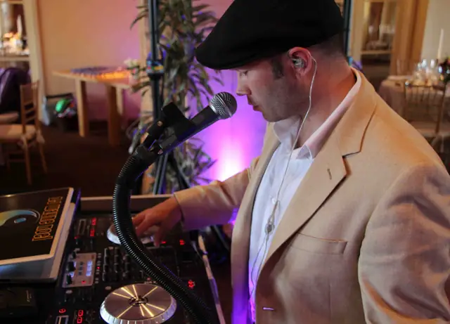 DJ Wayne Noel in a flat cap working a controller and speaking into a gooseneck mic at a wedding reception at Auberge du Soleil, Rutherford