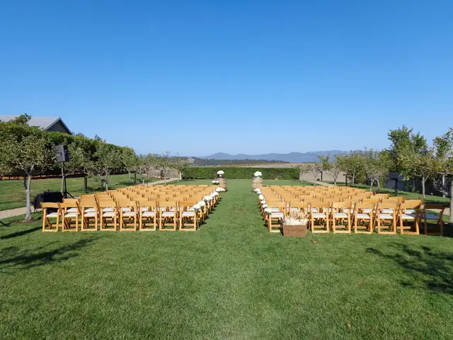 Outdoor ceremony lawn at Carneros Resort and Spa in Napa with wooden folding chairs either side of a grass aisle, orchard trees and one speaker on a stand at left