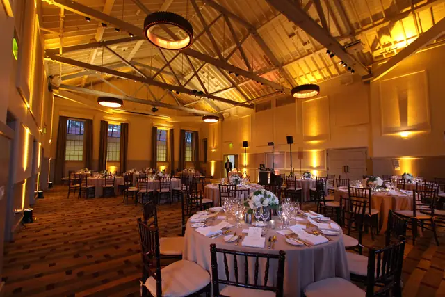 Amber uplighting across the trussed ballroom at Cavallo Point in Sausalito, set with round dinner tables and DJ Wayne Noel's booth and speakers at the far wall
