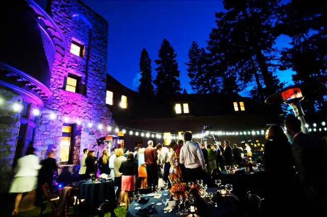 Guests mingling at dusk on the lawn at the Ehrman Mansion in Lake Tahoe, with the stone tower lit purple and string lights overhead
