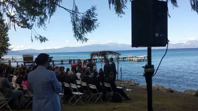 Lakeside wedding ceremony at Lake Tahoe with guests in white chairs, a flower arch by the pier and a DJ speaker on a stand in the foreground