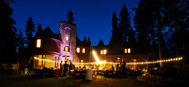 The Ehrman Mansion at night on Lake Tahoe, its stone tower lit purple and a line of string lights over the wedding reception lawn
