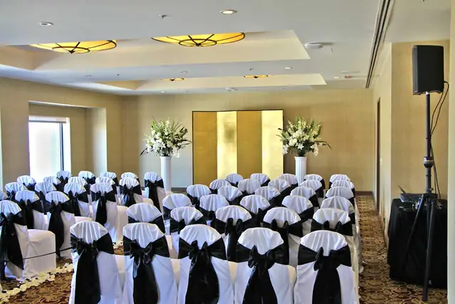 Wedding ceremony chairs with black sashes facing a gold screen and flowers at Hotel Nikko San Francisco, with a DJ speaker on a stand at right