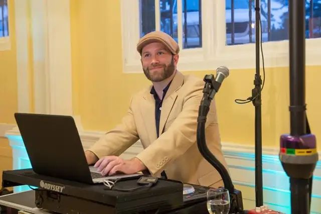 DJ Wayne Noel in a tan cap and blazer at his laptop and mixer beside a microphone, with tall windows and yellow walls of the Hotel Petaluma ballroom behind him