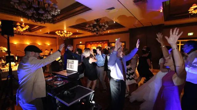Wedding guests with their hands in the air on the ballroom dance floor at The Inn at Spanish Bay in Pebble Beach, with the DJ raising an arm at the decks