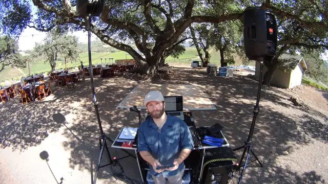 DJ Wayne Noel at his setup with two speakers on stands under a large oak tree at Kunde Family Winery, with reception tables and vineyard hills behind