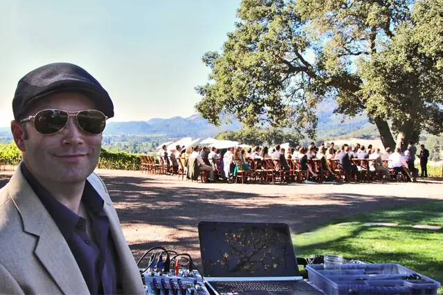 DJ Wayne Noel at his mixer and laptop with an outdoor wedding ceremony and valley view behind him at Kunde Family Winery