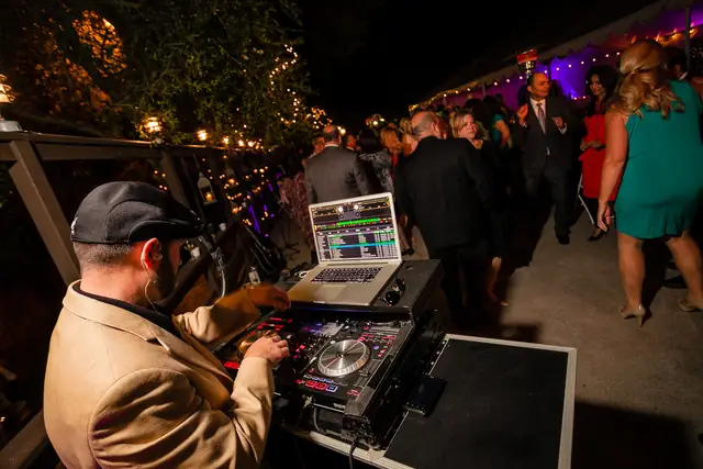 DJ Wayne Noel at his controller and laptop on a wooden deck at Little Hills in San Ramon, with a string-lit tent and guests dancing behind him