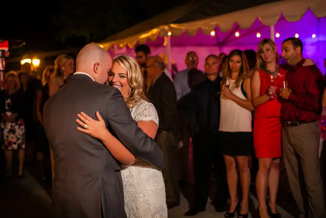A bride smiles over her groom's shoulder during their first dance on the patio at Little Hills in San Ramon, guests watching beside the uplit tent