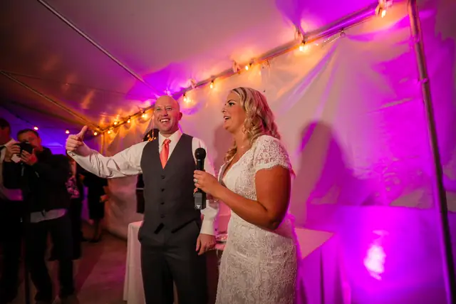 A bride and groom welcome their guests on the microphone inside the magenta-lit tent at Little Hills in San Ramon