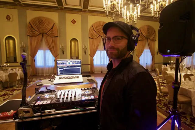 DJ Wayne Noel behind his controller and laptop in the Marines' Memorial Club ballroom, with chandeliers and draped windows behind him