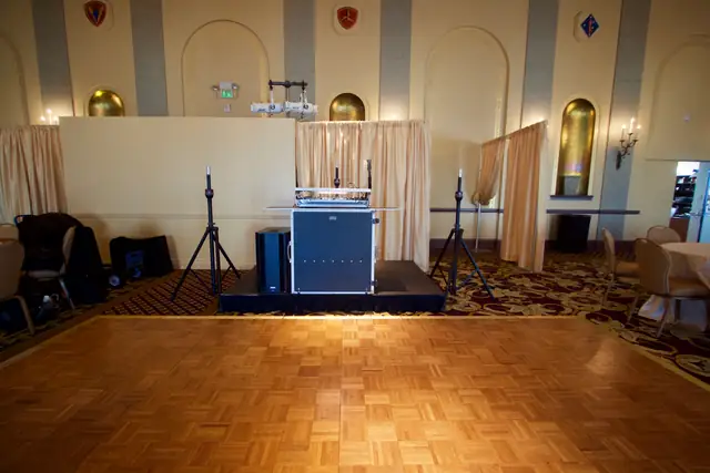 DJ setup on a skirted riser beside the parquet dance floor in the Marines' Memorial Club ballroom, with arched gold alcoves and Marine Corps insignia on the walls