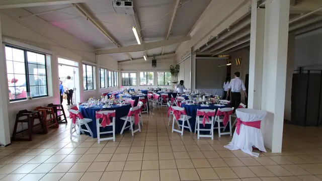 Reception room at Mavericks House set with navy round tables and pink-sashed chairs for a wedding, windows along the far wall