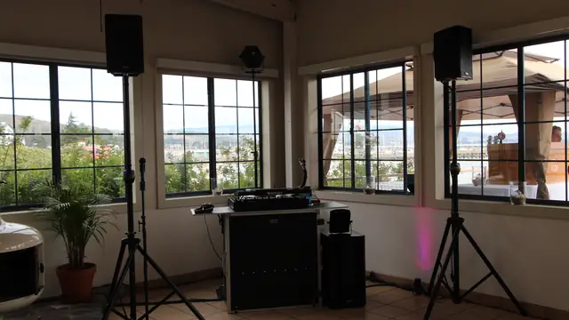 DJ booth and two speakers on stands set in front of harbor-view windows at Mavericks House