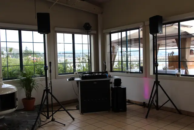 DJ booth with two speakers on stands and a subwoofer set up beside the windows at Mavericks House, with the harbor visible outside