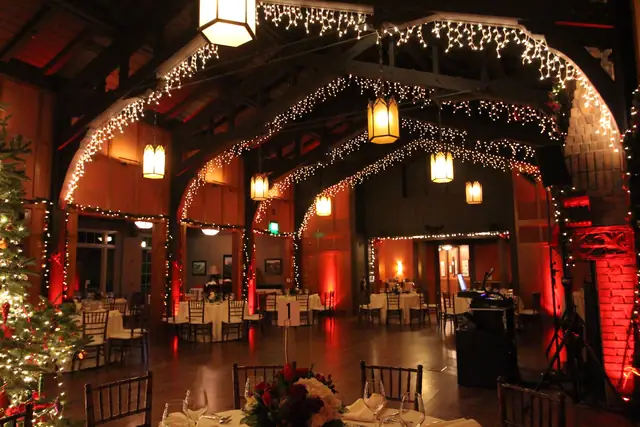 Timber-framed ballroom at the Meadow Club with arched trusses, hanging lanterns, set round tables and red uplighting, with a DJ booth at the right