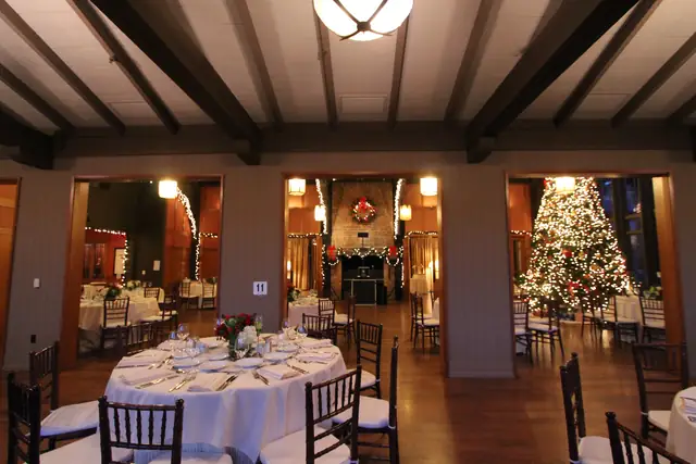 Meadow Club dining room set with round tables, with a DJ booth set up in the fireplace alcove beyond and a lit Christmas tree