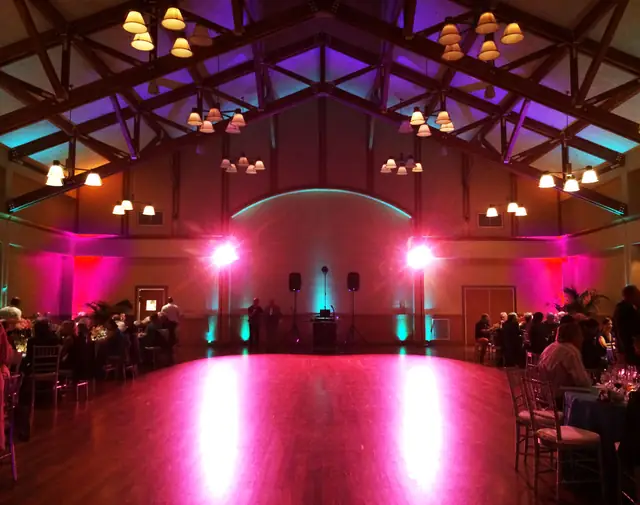 Vaulted timber-truss ballroom at the Mill Valley Community Center lit in pink and teal with a DJ setup at the far wall