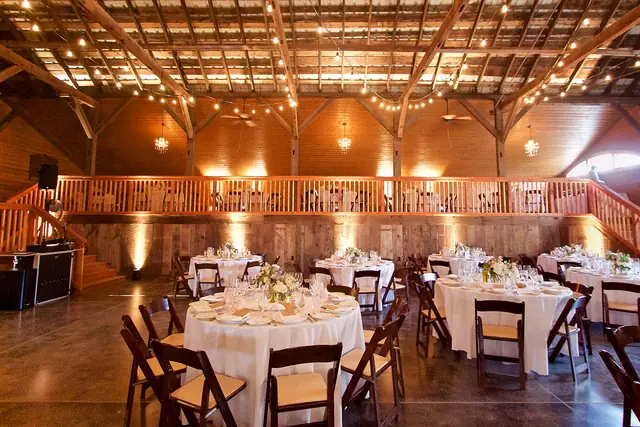 Reception room inside the barn at Olympia's Valley Estate with round tables, a wooden balcony, string lights and warm uplighting, with Wayne's DJ booth at the foot of the stairs