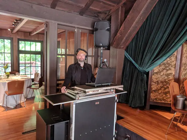 DJ Wayne Noel at his booth on the stage at the Mill Valley Outdoor Art Club, with a green velvet curtain, wood beams and set dinner tables behind him