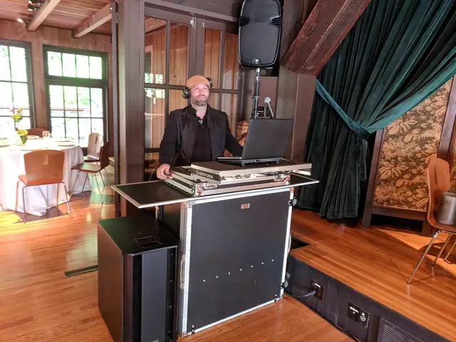 DJ Wayne Noel at his booth beside the green velvet stage curtain inside the Mill Valley Outdoor Art Club, with the dining room set behind him