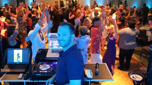 DJ Wayne Noel smiling behind his controller and laptops while guests dance with their arms raised in a ballroom at Seascape Beach Resort