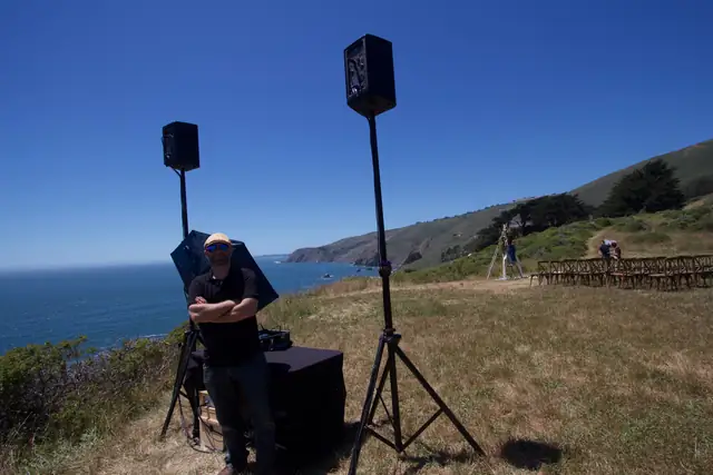 DJ Wayne Noel beside his speakers on the bluff at Slide Ranch with the coast and ceremony chairs behind