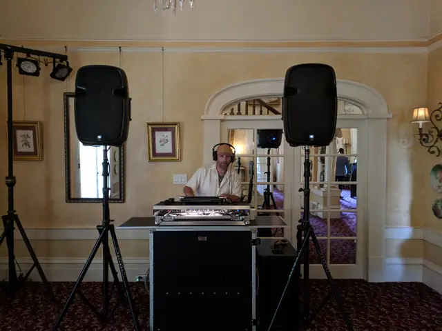 DJ Wayne Noel at his decks in a parlor at The Madrona, with two speakers on stands and stage lights on a bar beside the arched French doors
