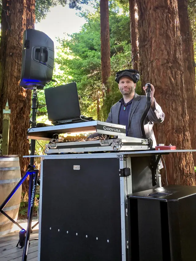 DJ Wayne Noel at his booth among the redwoods at The Secret Gardens in Bodega Bay, with a speaker on a tripod and a wine barrel beside the deck