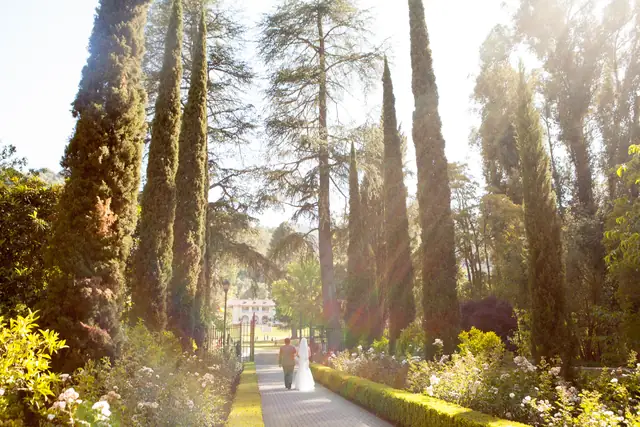 Bride and escort walking down the cypress-lined rose garden path at Villa Montalvo toward the villa in warm backlight