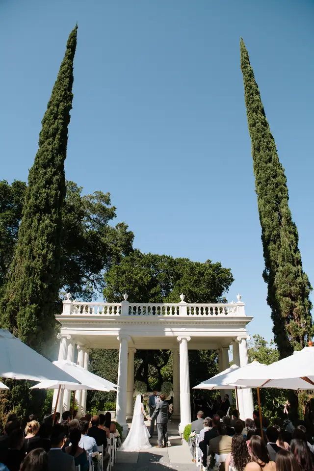 Wedding ceremony at the white colonnade pavilion in the rose garden at Villa Montalvo, framed by two tall cypress trees