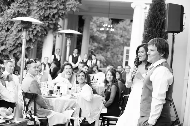 A bride speaks into a wireless microphone beside the groom during dinner on the terrace at Villa Montalvo in Saratoga, with a speaker on a stand nearby